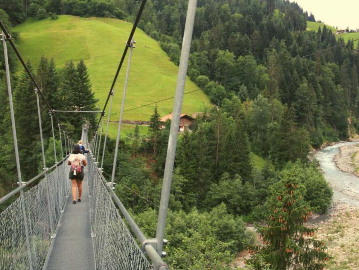young women walking across suspension bridge with mountains and river below