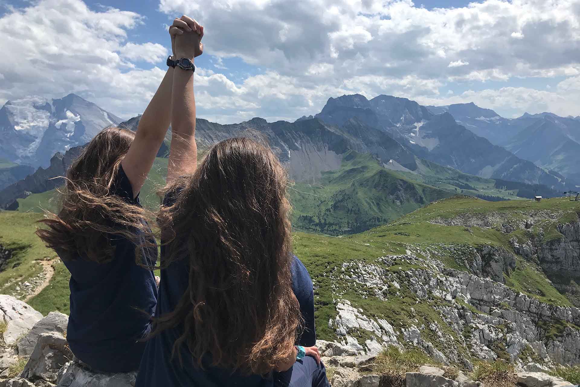 Two girls holding hands up high in a celebratory way while looking out over a valley from a high mountaintop
