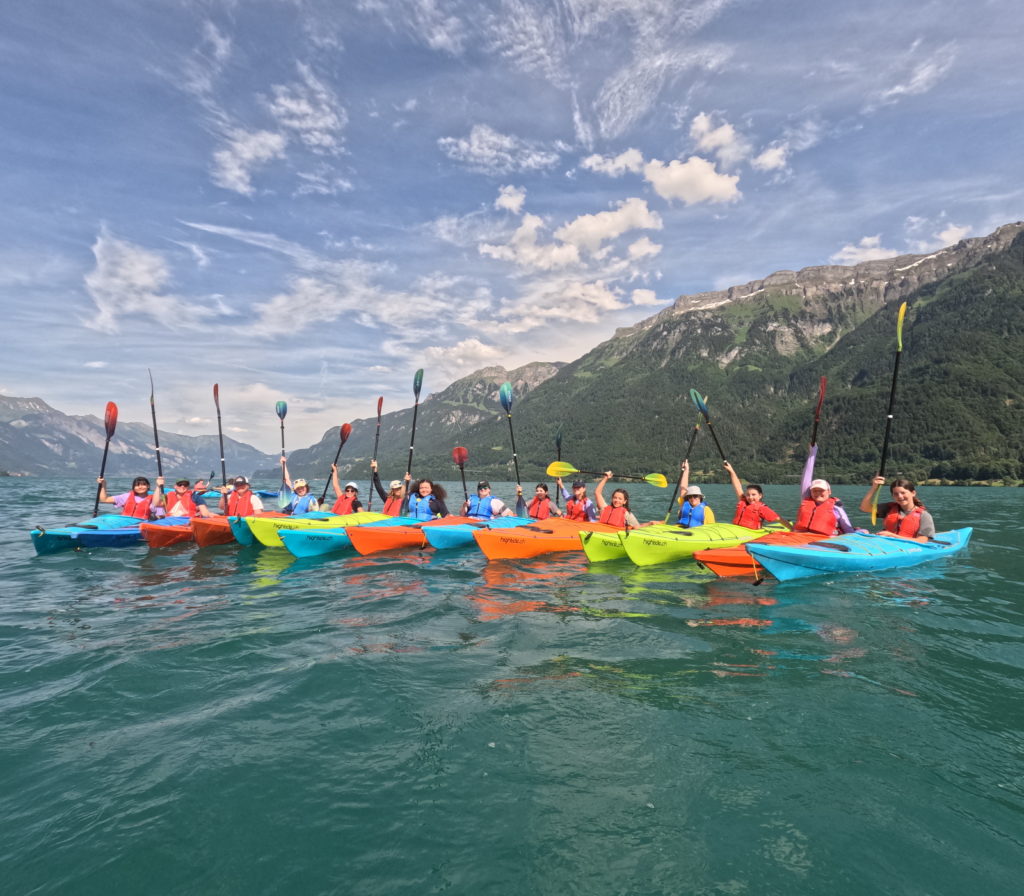 A group of young women on a lake in colourful kayaks holding their paddles in the air. Mountains in the background.