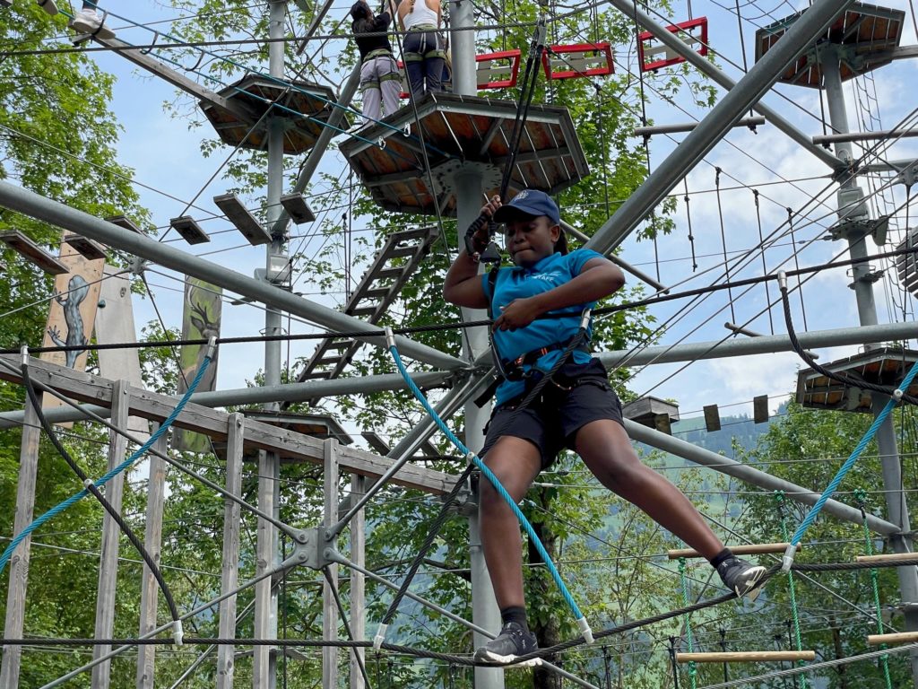 A young black woman climbing across netting on a high ropes course. Her face shows concentration.